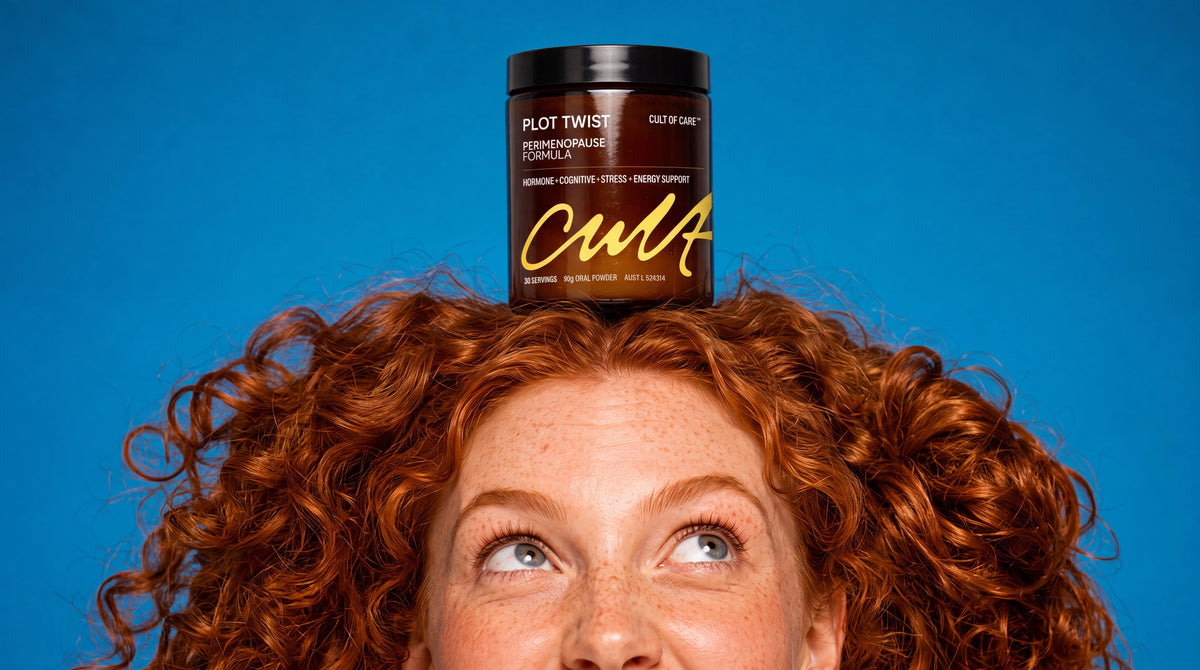 A person with curly red hair balances a black jar of Cult of Care Plot Twist Perimenopause Formula on her head against a solid blue background.