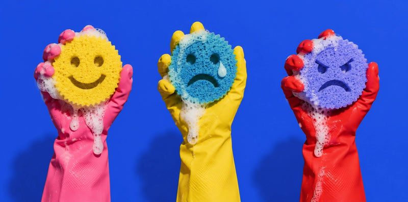 Three gloved hands holding colorful textured sponge scrubbers with facial expressions, set against a solid blue background.