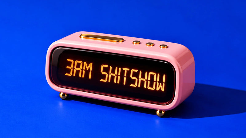 A pink retro-style digital alarm clock with gold accents and a black display screen showing the text '3AM SHITSHOW' in glowing orange letters, set against a solid blue background.
