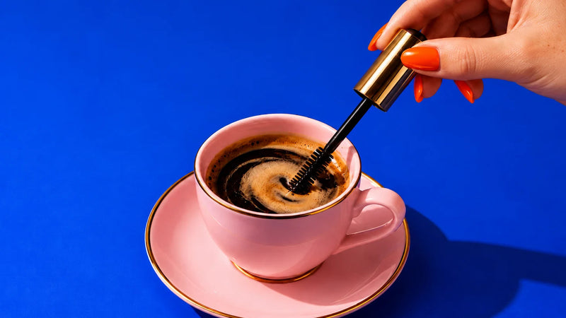 A hand with orange nail polish holds a black and gold mascara and is stirring the coffee in a pink cup on a bright blue background.