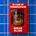 A jar of Cult of Care Plot Twist Perimenopause Formula sits inside a red fire alarm box mounted on a blue tiled wall. The red box has white text reading 'IN CASE OF PERIMENOPAUS' at the top and 'BREAK GLASS' at the bottom.