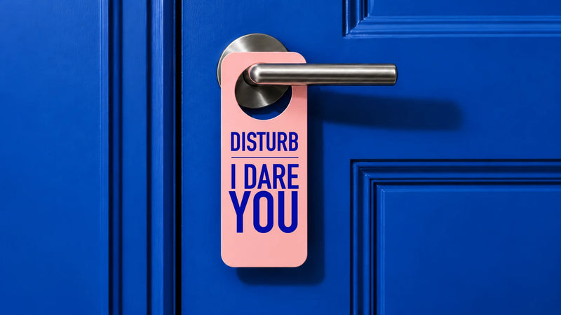 A pink hanger tag with the text 'DISTURB I DARE YOU' in blue lettering, hanging from a silver metal doorknob on a bright blue paneled door.