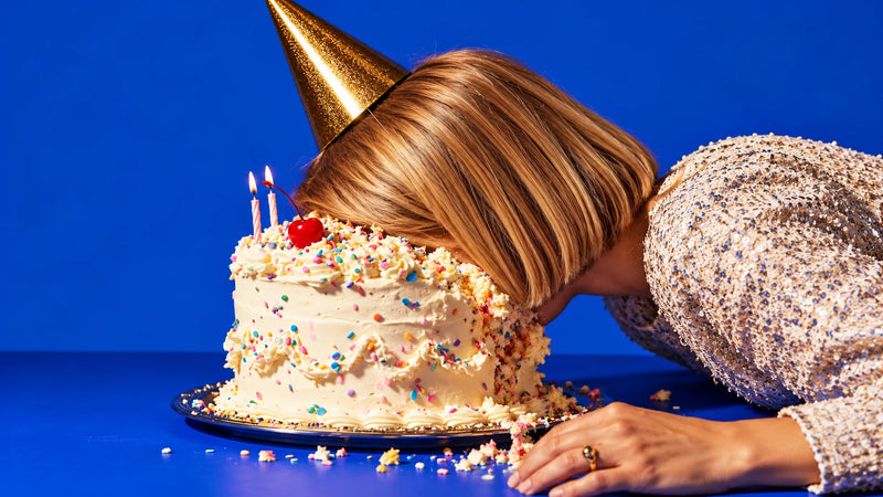 A woman with blonde hair wearing a gold party with her face squashed into a frosted birthday cake with sprinkles and lit candles, set against a solid blue background.