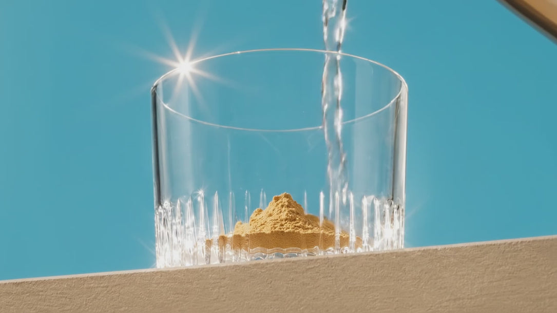 A video of water pouring into a glass of with Plot Twist powder in the bottom. The water turns to a beautiful golden orange colour as the water and powder mix. Styled against a sky blue background with light refracting on the glass and through the liquid
